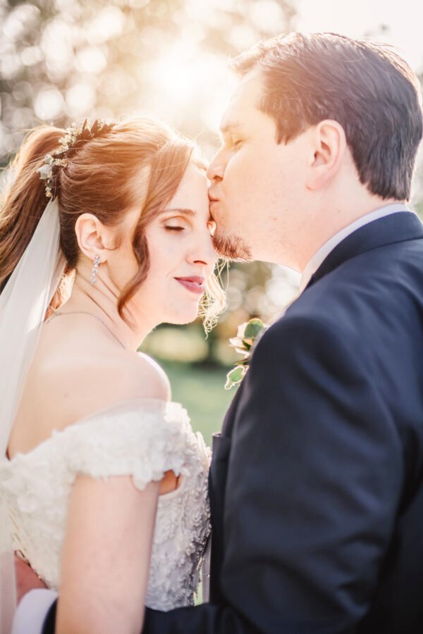 Tendresse au crépuscule : un moment romantique au coucher du soleil Un mari embrasse tendrement sa femme sur le front, tandis que la mariée regarde l’objectif avec douceur. Le coucher de soleil les enveloppe d’une lumière dorée et romantique