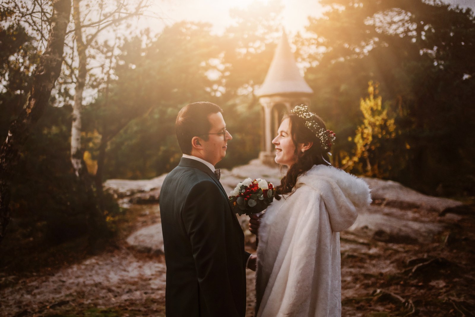 Couple de mariés marchant main dans la main sous les arbres dans une forêt d’automne, baignée par une lumière dorée de novembre.