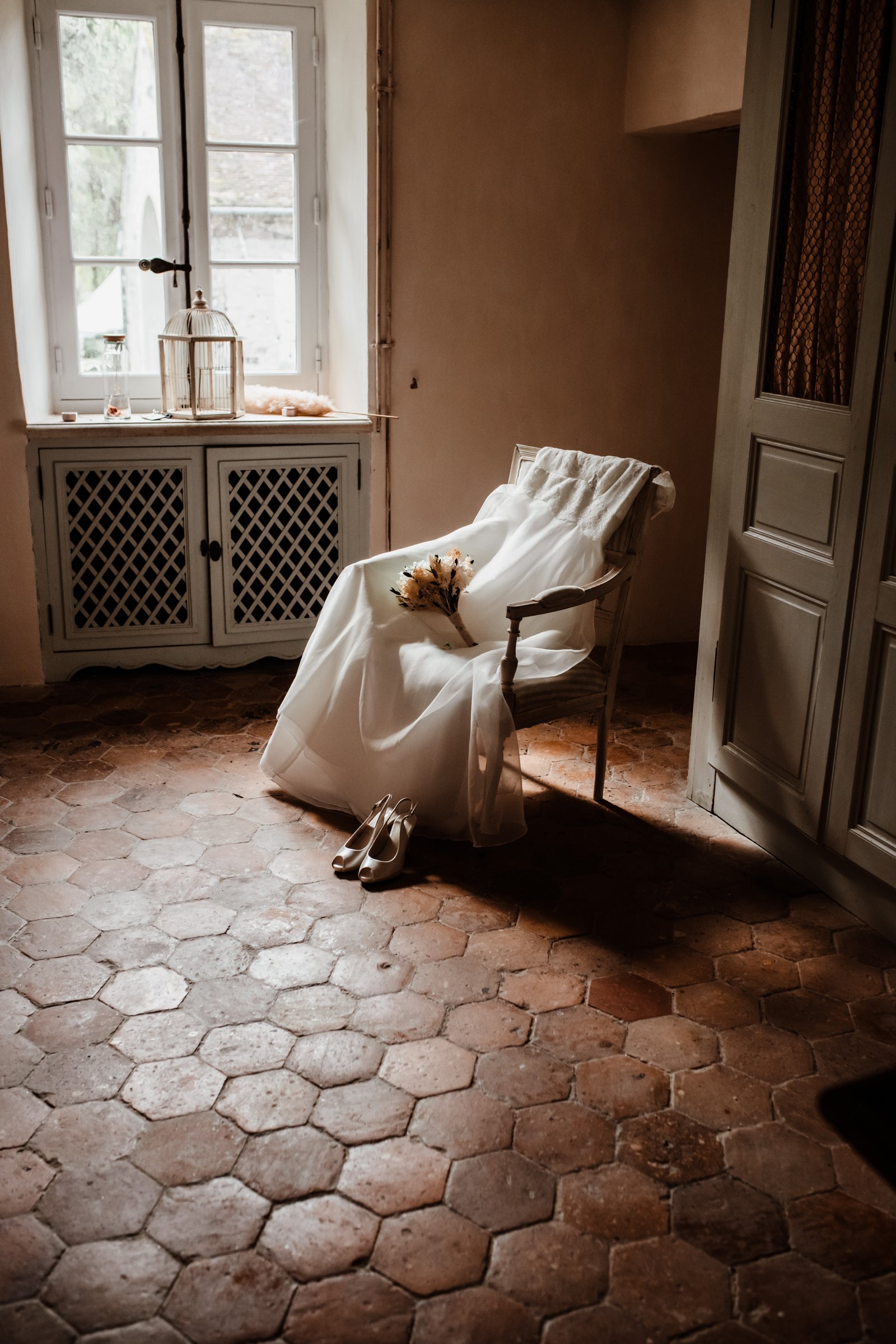 La délicatesse d’une robe de mariée baignée de lumière Robe de mariée délicatement posée sur une chaise avec un bouquet de fleurs, illuminés par un rayon de soleil doux et chaleureux.