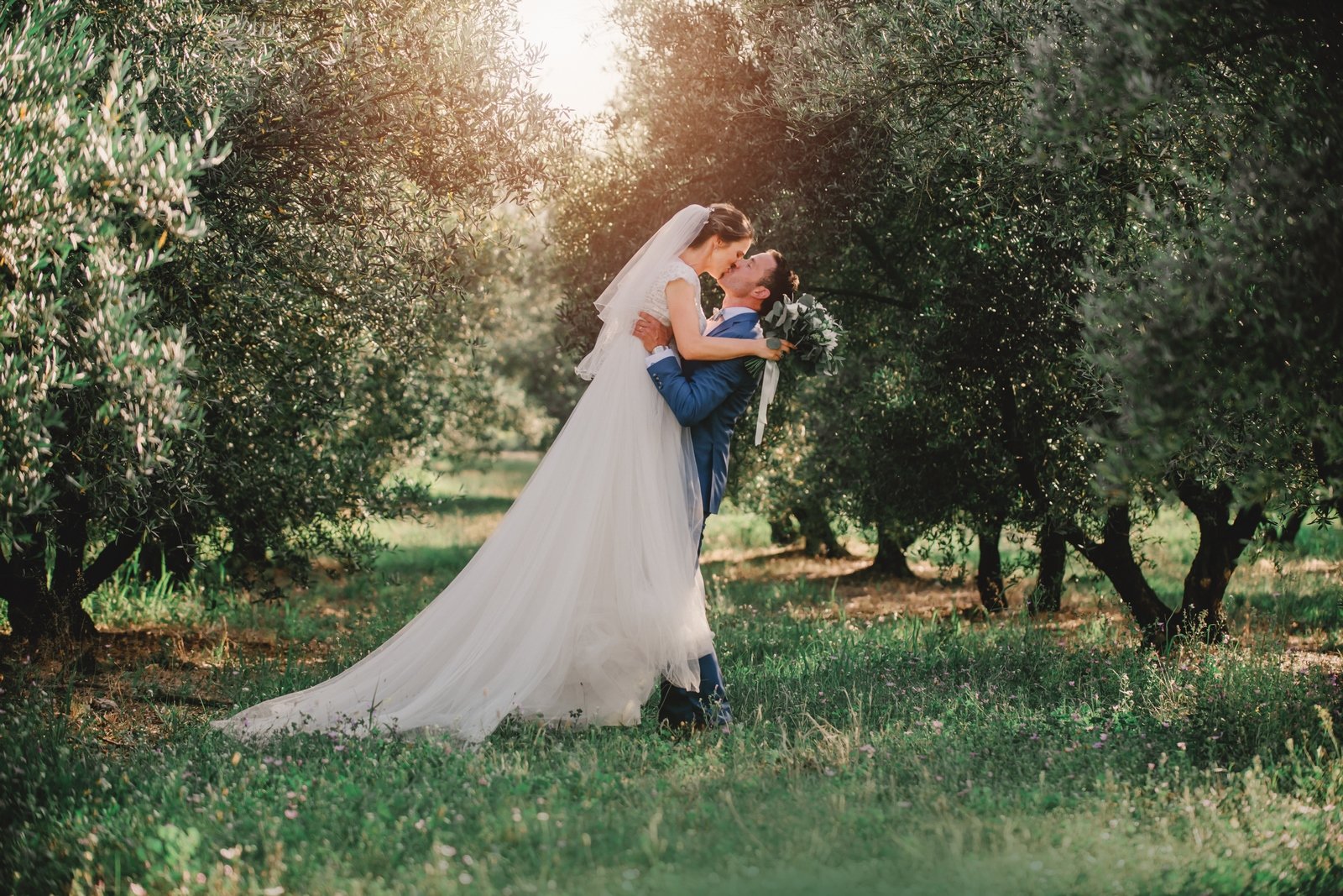 Un baiser romantique au cœur d’une oliveraie provençale Couple de mariés s’embrassant dans les allées d’une oliveraie en Provence, entourés d’oliviers et baignés par une lumière douce et dorée.