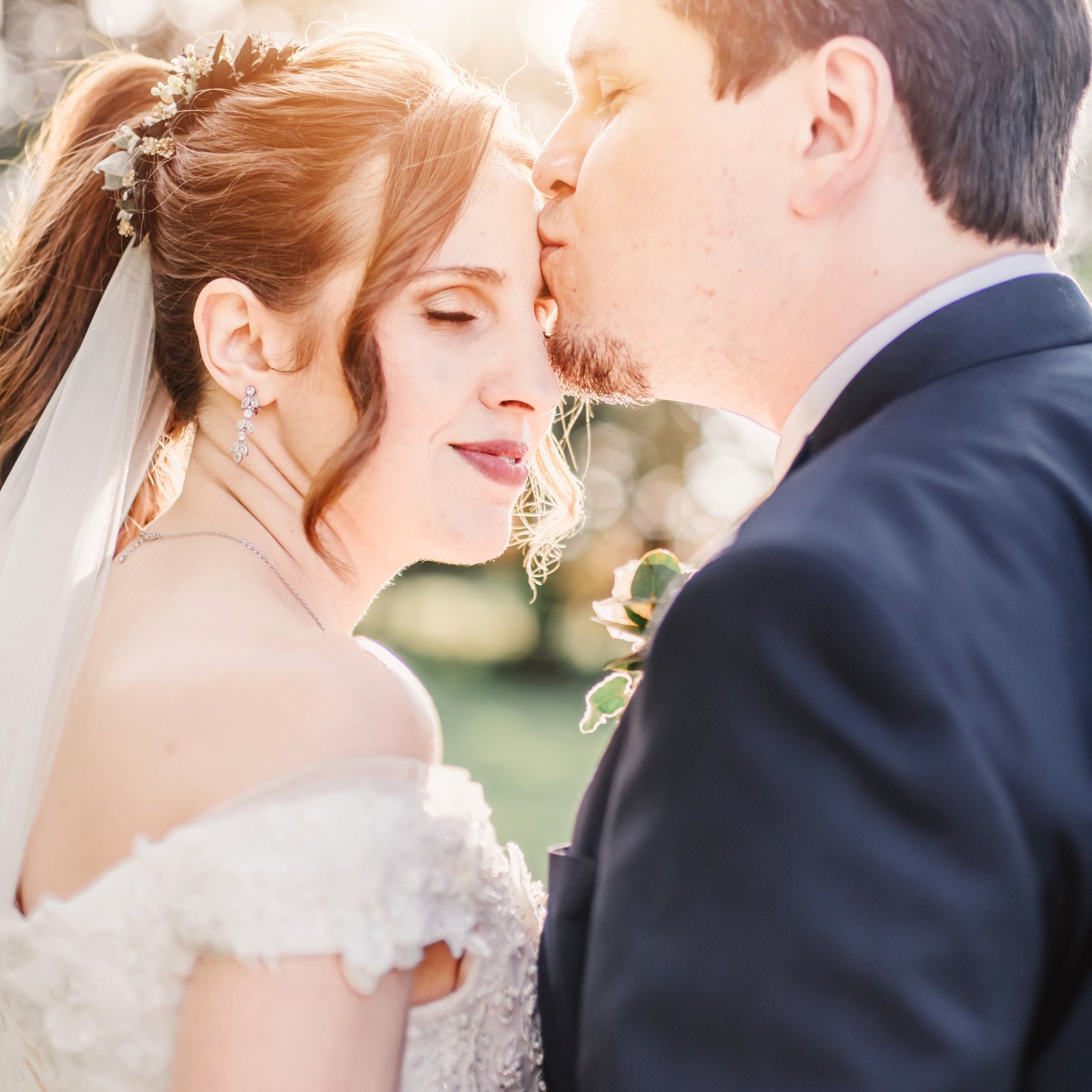 Un mari embrasse tendrement sa femme sur le front, tandis que la mariée regarde l’objectif avec douceur. Le coucher de soleil les enveloppe d’une lumière dorée et romantique